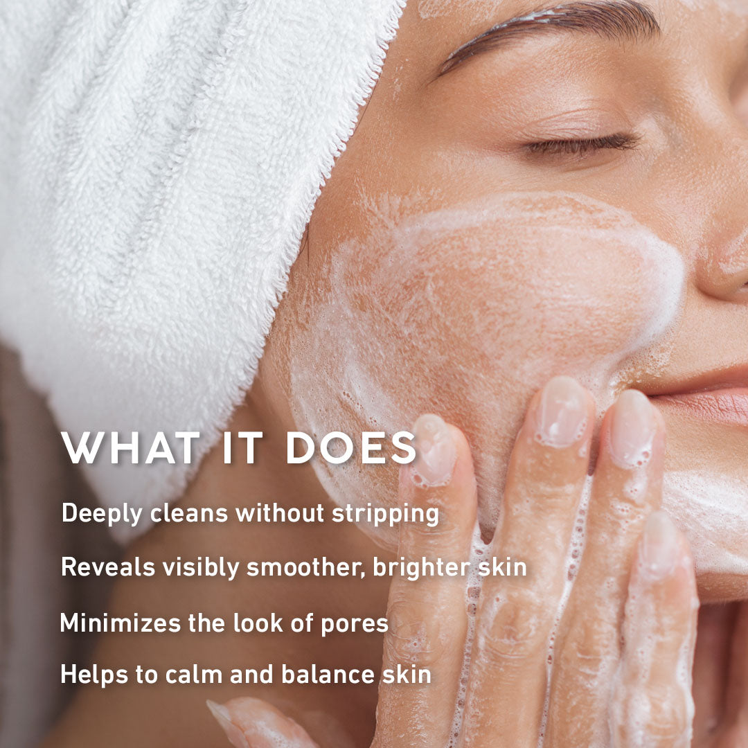 Close-up of a woman with a white towel wrapped around her hair while applying or washing her face with foaming skincare product. White text on the left reads 'WHAT IT DOES' followed by four benefits about deep cleansing, smoother skin, pore minimization, and skin balance.