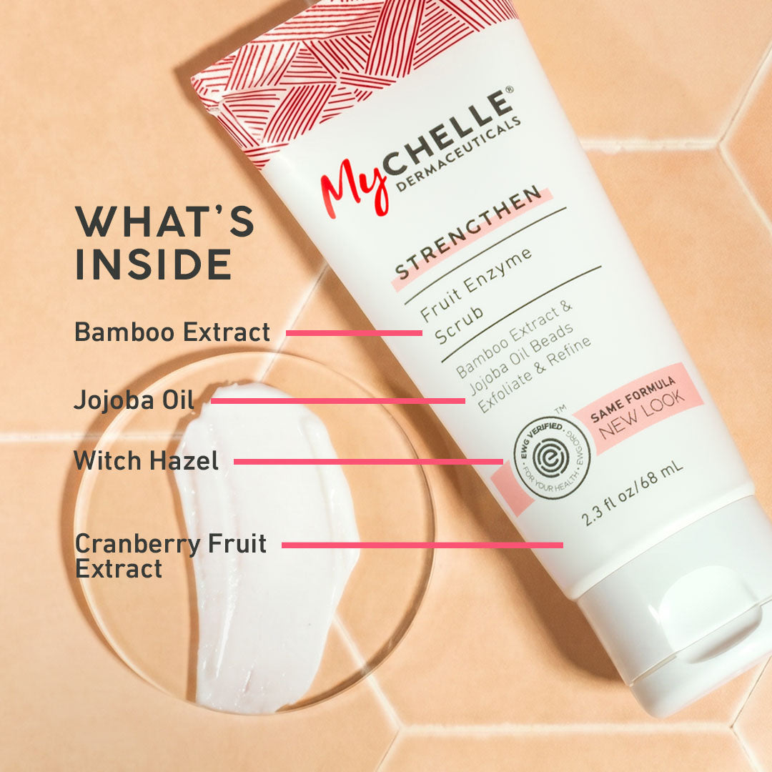 MyCHELLE Dermaceuticals Fruit Enzyme Scrub in a 2.3 fl oz squeeze tube, shown diagonally on a beige hexagonal background with a smear of white scrub and text listing ingredients: Bamboo Extract, Jojoba Oil, Witch Hazel, and Cranberry Fruit Extract.